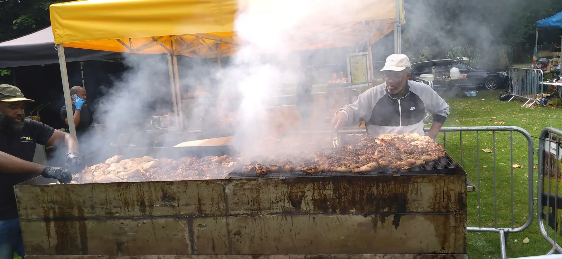 Two Black cooks working over a large smoking jerk grill at Manchester Caribbean Carnival