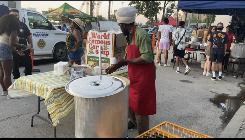 A Caribbean diaspora food vendor serving corn soup from a large street pot