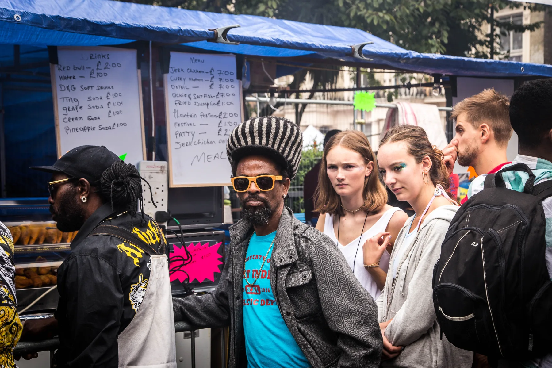 Carnival-goers queueing beside a handwritten Caribbean food menu at Notting Hill Carnival