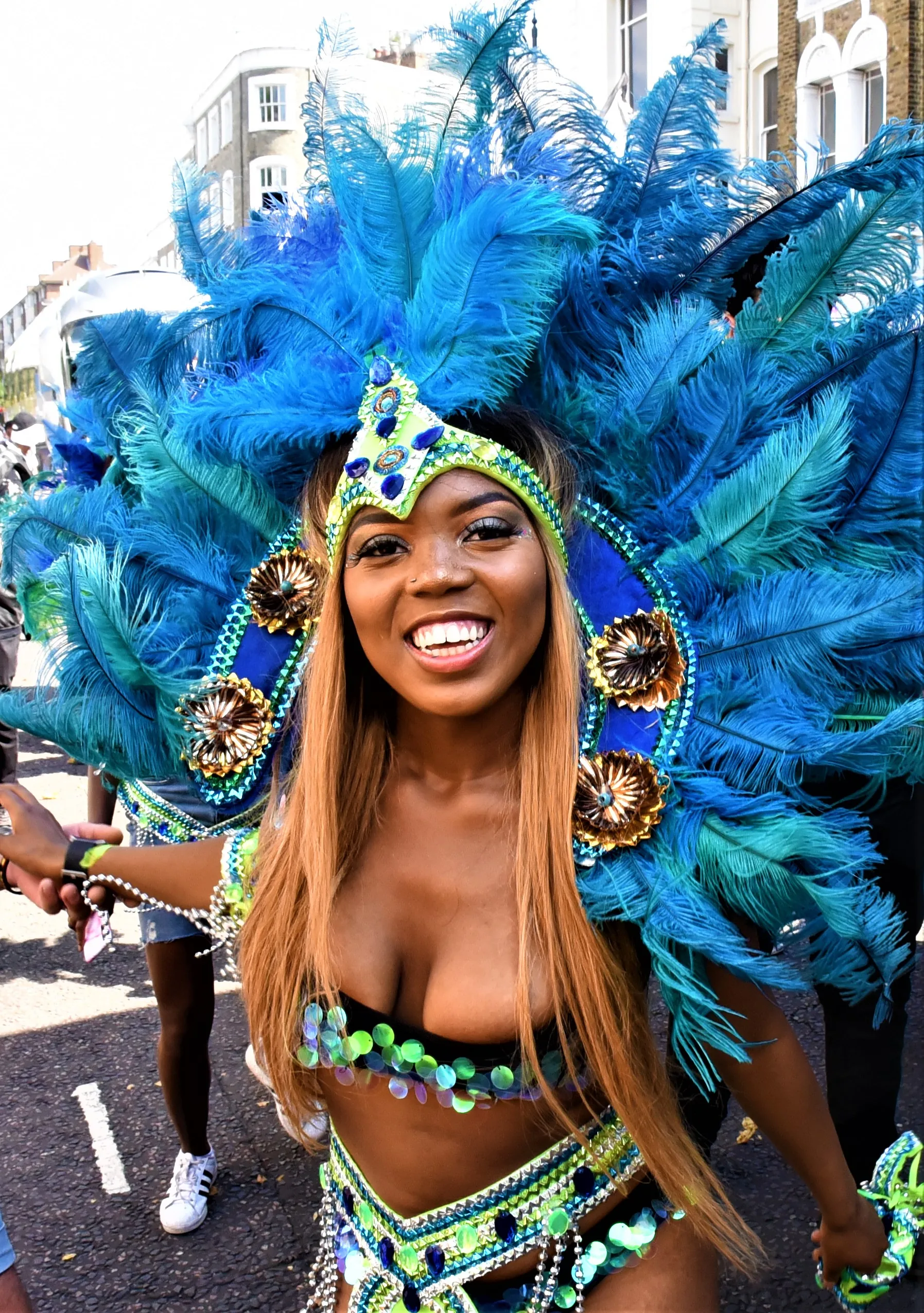 A smiling Black masquerader in a blue feathered costume playing mas at Notting Hill Carnival