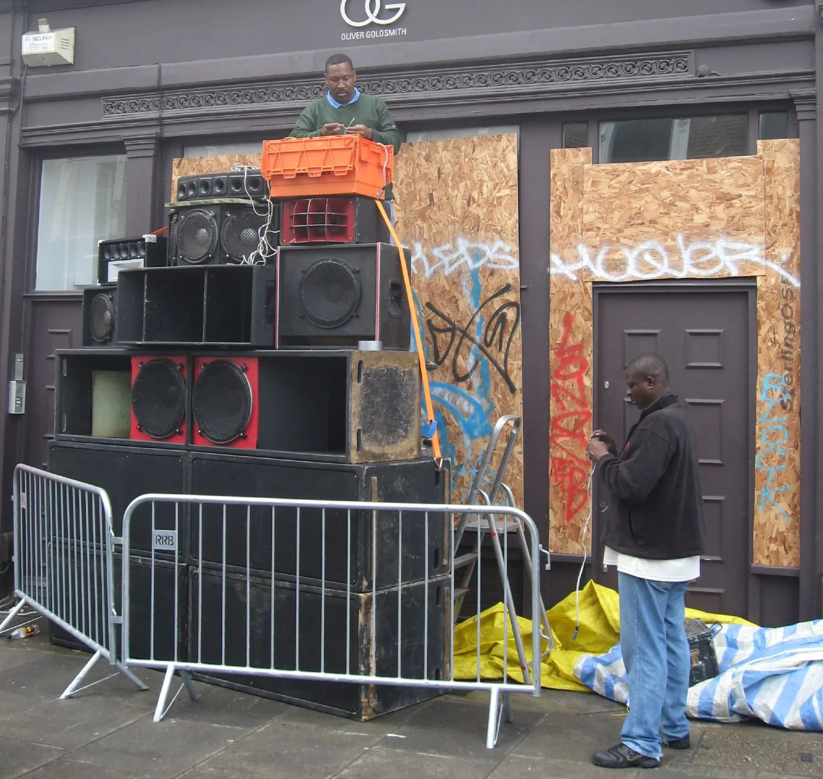 Two men setting up a tall, hand-built sound system on a west London street before Notting Hill Carnival
