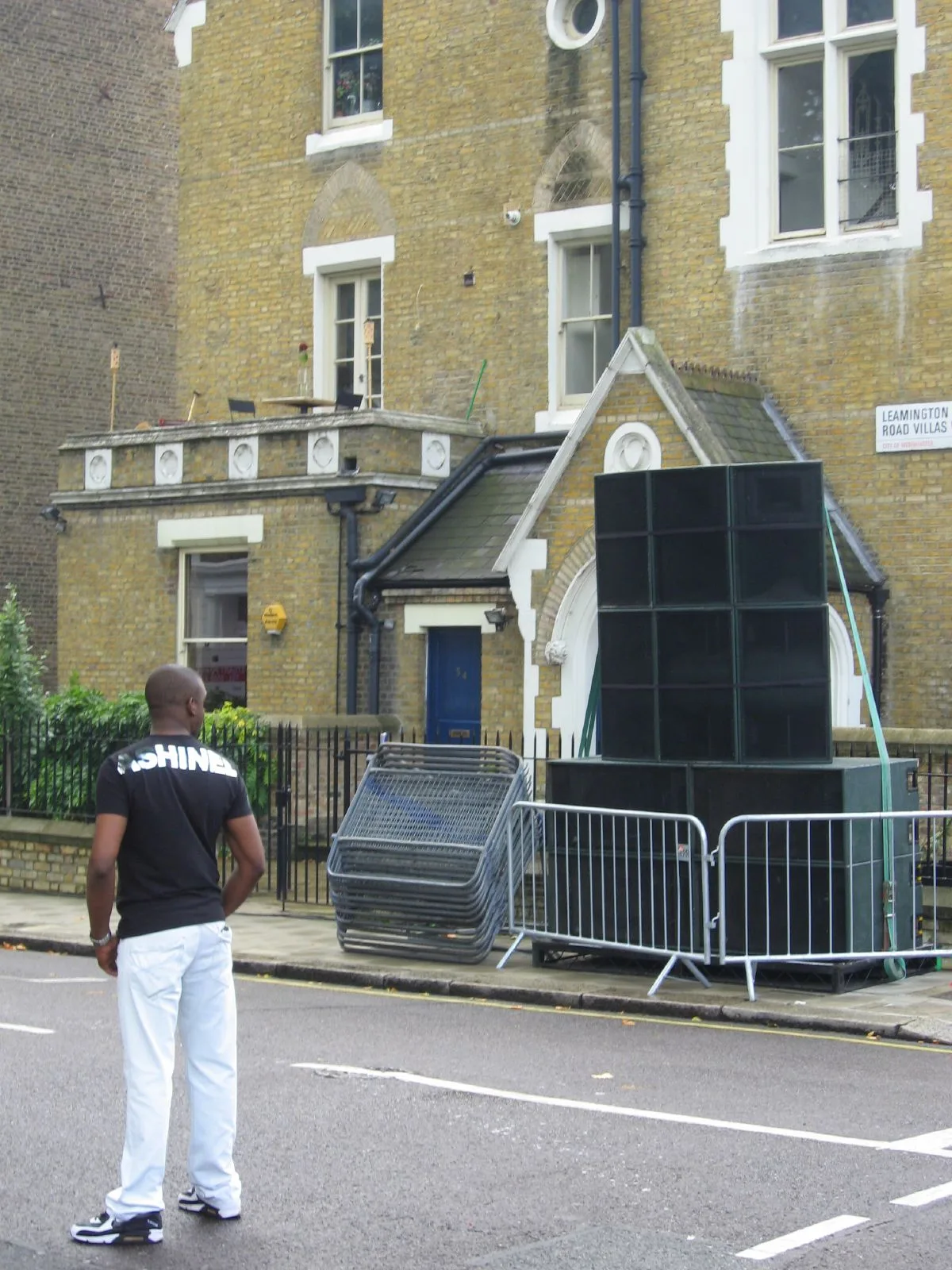 A Black man looks across Leamington Road Villas at a tall static sound-system stack behind barriers