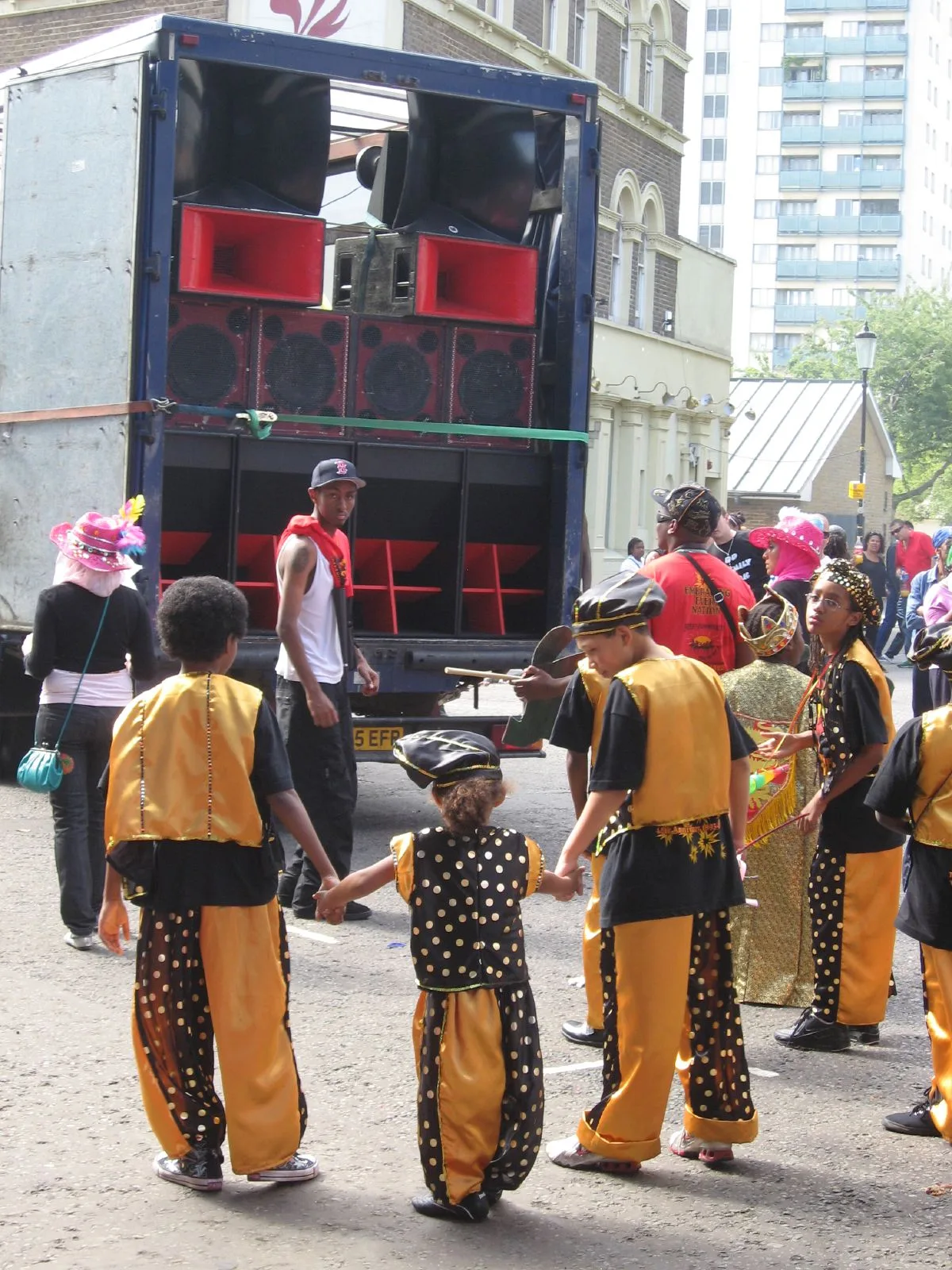A red and black sound system fills the back of a truck while a children's mas band passes on the street